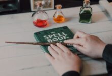 Close-up of hands with spell book, wand, and colorful potions on a table.