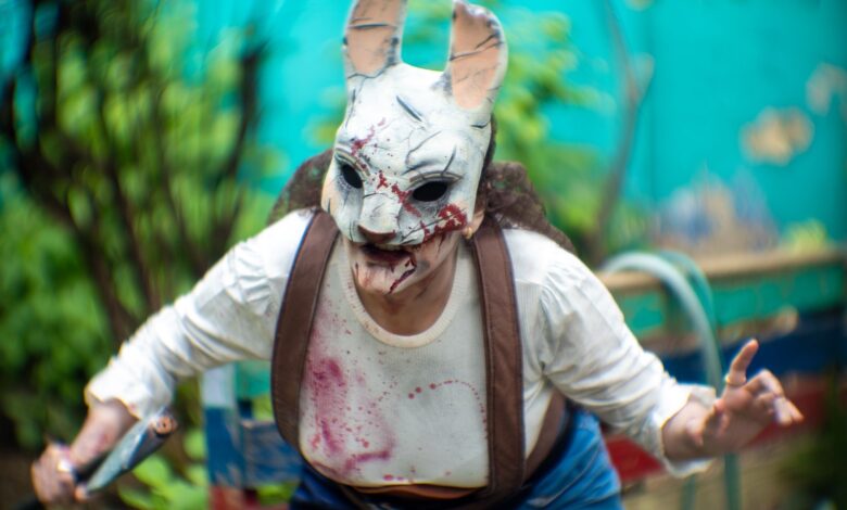 A person in a terrifying rabbit mask performs horror cosplay outdoors amid vibrant greenery.