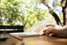 A hand flipping through a sketchbook with colorful drawings in a lush outdoor setting.