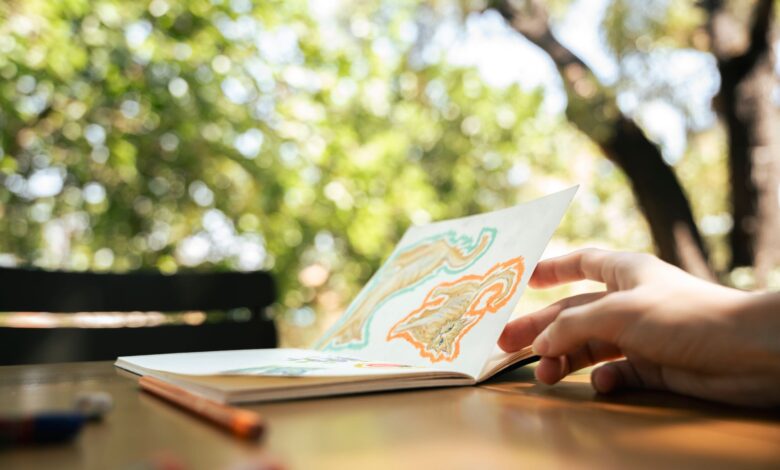 A hand flipping through a sketchbook with colorful drawings in a lush outdoor setting.