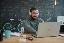 A young adult with headphones works on a laptop in a modern office with a chalkboard wall.