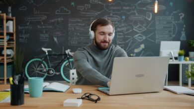 A young adult with headphones works on a laptop in a modern office with a chalkboard wall.