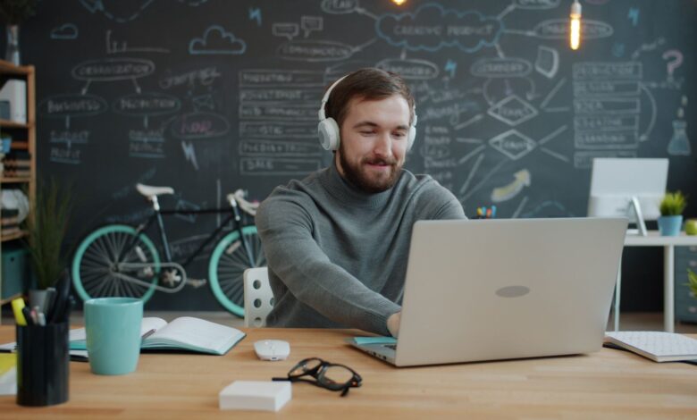 A young adult with headphones works on a laptop in a modern office with a chalkboard wall.