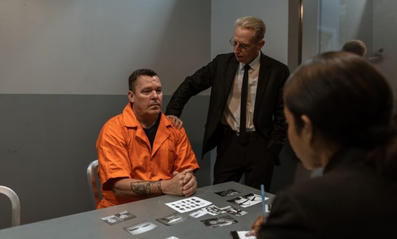 Prisoner in orange jumpsuit interviewed by detectives in a tense interrogation room scene.