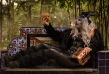 Gothic figure in costume holding a skull, seated on a decorated bench in a forested area.