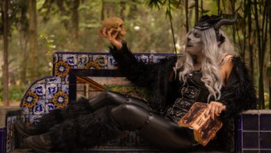 Gothic figure in costume holding a skull, seated on a decorated bench in a forested area.
