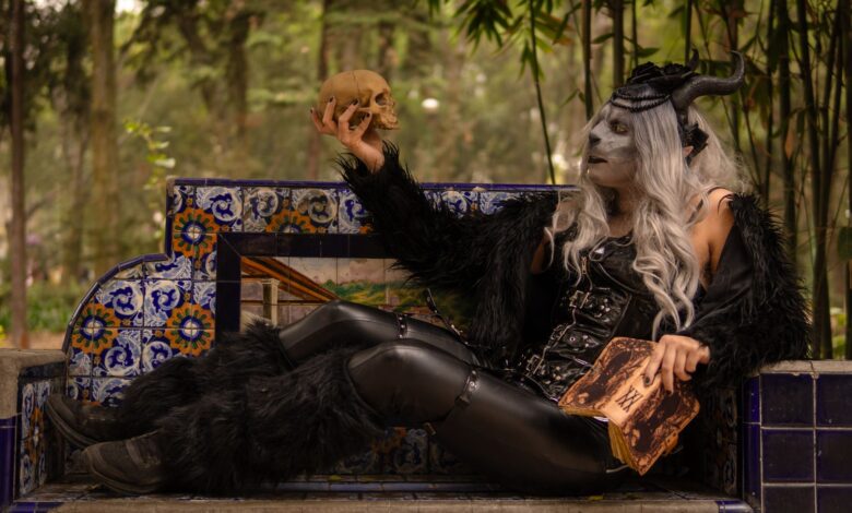 Gothic figure in costume holding a skull, seated on a decorated bench in a forested area.