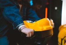 Hand holding handlebar of a yellow arcade racing game machine in a dimly lit setting.