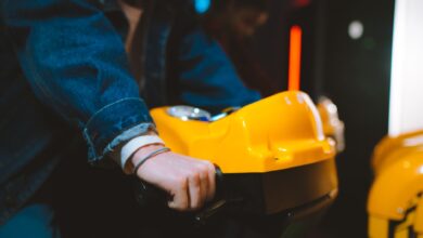 Hand holding handlebar of a yellow arcade racing game machine in a dimly lit setting.
