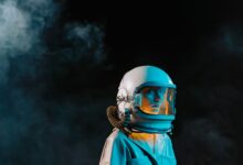 A dramatic portrait of a woman wearing a spacesuit and helmet in a dark, smoky setting.