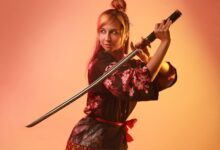 A woman in colorful cosplay posing with a samurai sword in a studio setting.