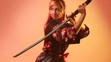 A woman in colorful cosplay posing with a samurai sword in a studio setting.