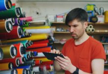 Man using a digital tablet in a print shop with colorful rolls of paper.
