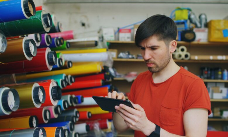 Man using a digital tablet in a print shop with colorful rolls of paper.