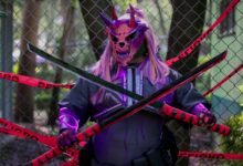 A person in a demon mask holds swords in front of a fence marked with warning tape.