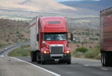 Red semi-truck driving through scenic highway in a mountainous landscape.