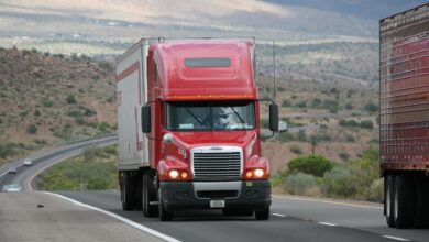 Red semi-truck driving through scenic highway in a mountainous landscape.