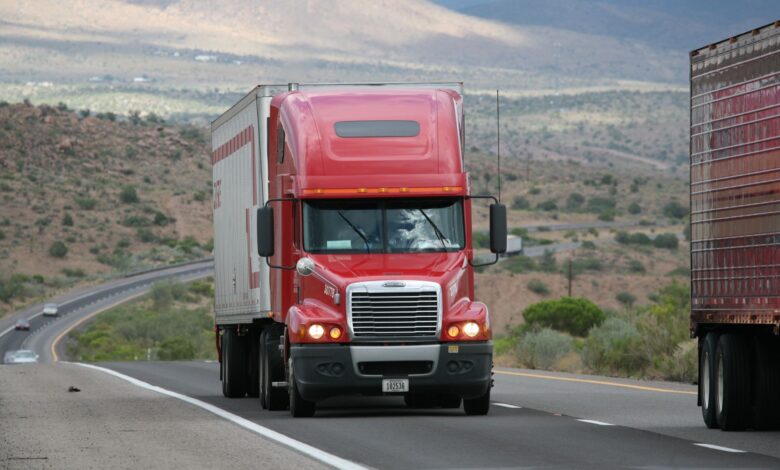 Red semi-truck driving through scenic highway in a mountainous landscape.