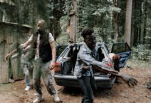 A group of zombies walking near an abandoned car in a creepy forest setting.