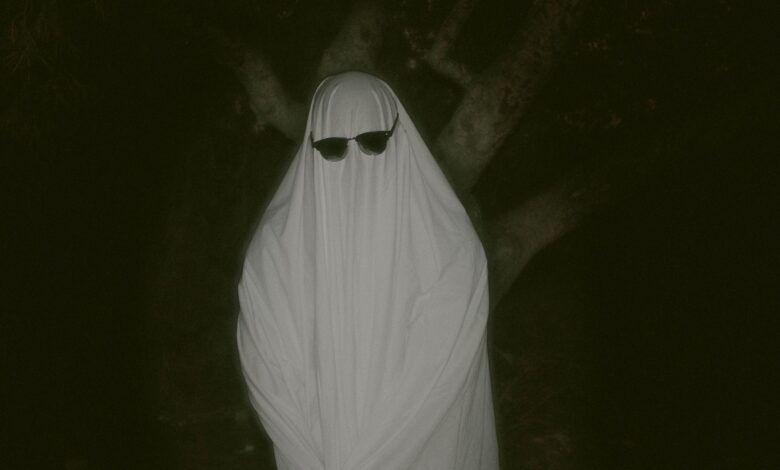 Mysterious figure dressed as a ghost wearing sunglasses in a dark forest setting.