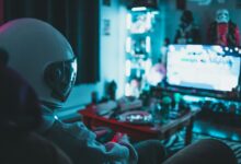 Side view of unrecognizable person in virtual reality helmet sitting on sofa and playing with gamepad in dark room