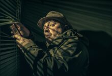 A man in a hat suspiciously peering through blinds, holding a phone in shadowy light.