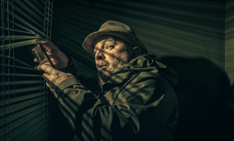 A man in a hat suspiciously peering through blinds, holding a phone in shadowy light.