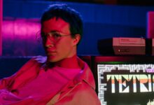 A teenager with eyeglasses in an 80s-style arcade displaying a Tetris game on screen.