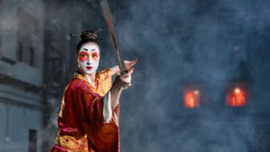 A woman in geisha makeup and traditional kimono poses with a sword amidst dramatic smoke effects.