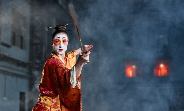 A woman in geisha makeup and traditional kimono poses with a sword amidst dramatic smoke effects.