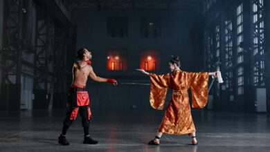 Two performers in martial arts costumes engage in a dramatic face-off inside a large industrial hall.