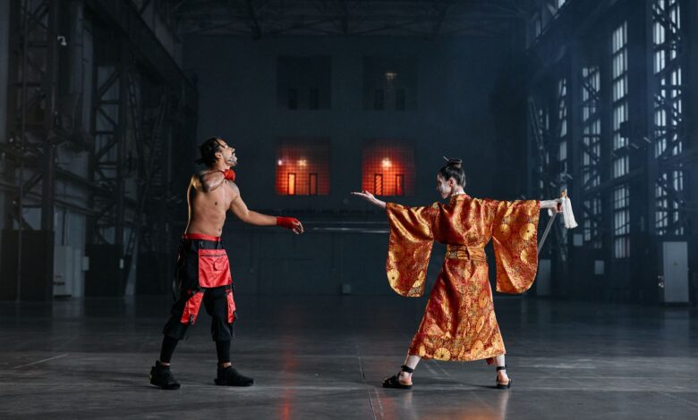 Two performers in martial arts costumes engage in a dramatic face-off inside a large industrial hall.