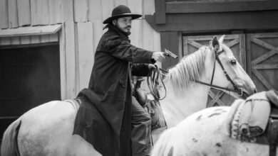 Black and white photo of a cowboy on horseback aiming a revolver in an old western setting.