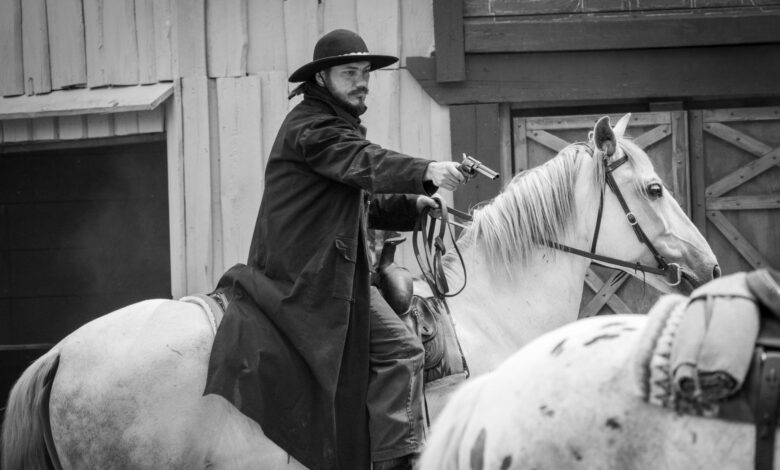 Black and white photo of a cowboy on horseback aiming a revolver in an old western setting.