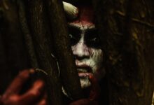 Scary demon-like face with stage makeup peeking through branches in a dark forest.