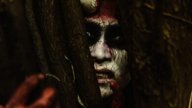 Scary demon-like face with stage makeup peeking through branches in a dark forest.