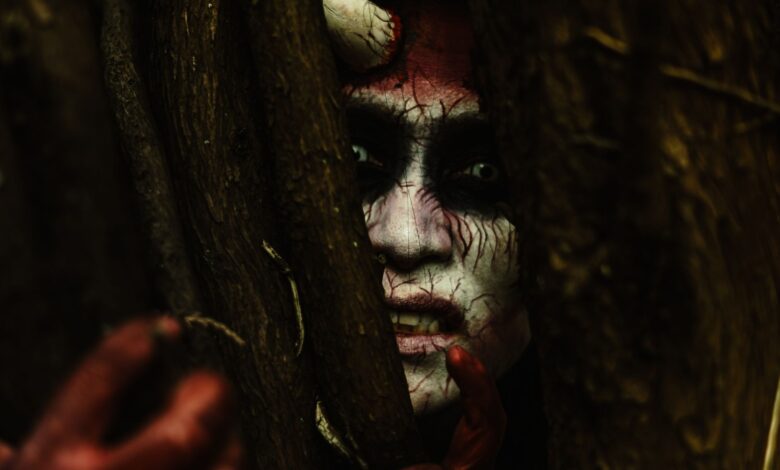 Scary demon-like face with stage makeup peeking through branches in a dark forest.