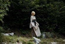 A cosplayer dressed as an elf in a fantasy costume stands in a lush, green forest.