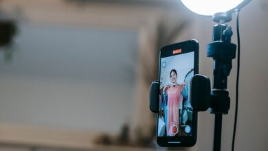 Anonymous female blogger standing in room and shooting vlog on smartphone with ring lamp while demonstrating pink dress in hands