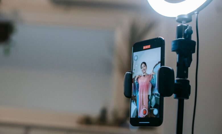 Anonymous female blogger standing in room and shooting vlog on smartphone with ring lamp while demonstrating pink dress in hands