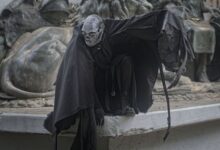 A mysterious figure in dark, theatrical costume crouches by a dramatic sculpture, enhancing a sense of mystery.