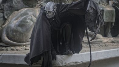 A mysterious figure in dark, theatrical costume crouches by a dramatic sculpture, enhancing a sense of mystery.