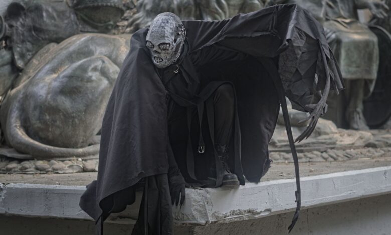 A mysterious figure in dark, theatrical costume crouches by a dramatic sculpture, enhancing a sense of mystery.