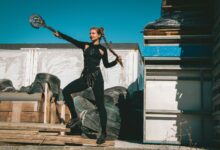 A woman dressed in a warrior costume poses outdoors with braided hair, holding a weapon.