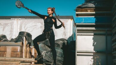 A woman dressed in a warrior costume poses outdoors with braided hair, holding a weapon.
