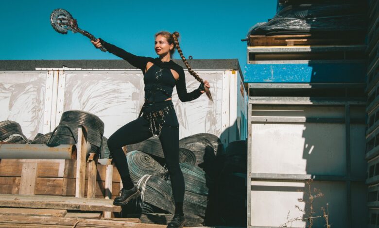 A woman dressed in a warrior costume poses outdoors with braided hair, holding a weapon.