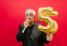 Senior man with a party horn and gold balloon, celebrating in a studio with a vibrant red background.