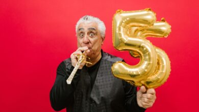 Senior man with a party horn and gold balloon, celebrating in a studio with a vibrant red background.