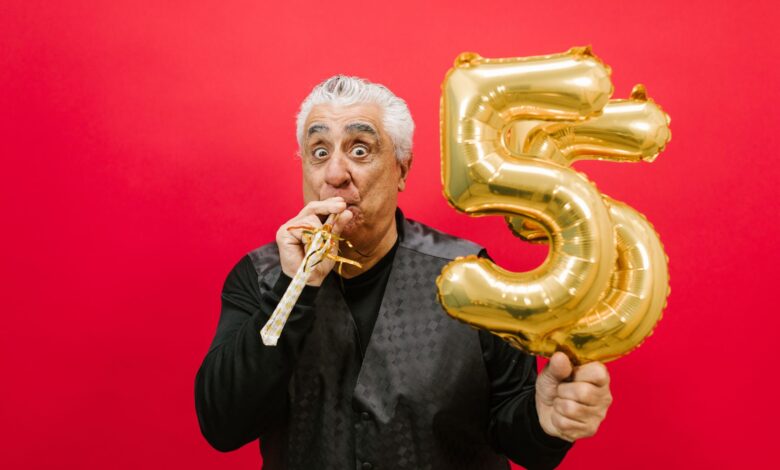 Senior man with a party horn and gold balloon, celebrating in a studio with a vibrant red background.
