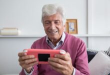 Elderly man smiling while playing on a handheld console indoors.